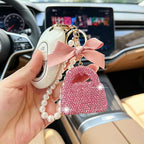 A hand holds a pink rhinestone handbag-shaped car keychain charm with a pearl strap and a pink satin bow, attached to a white car key fob, inside a car.
