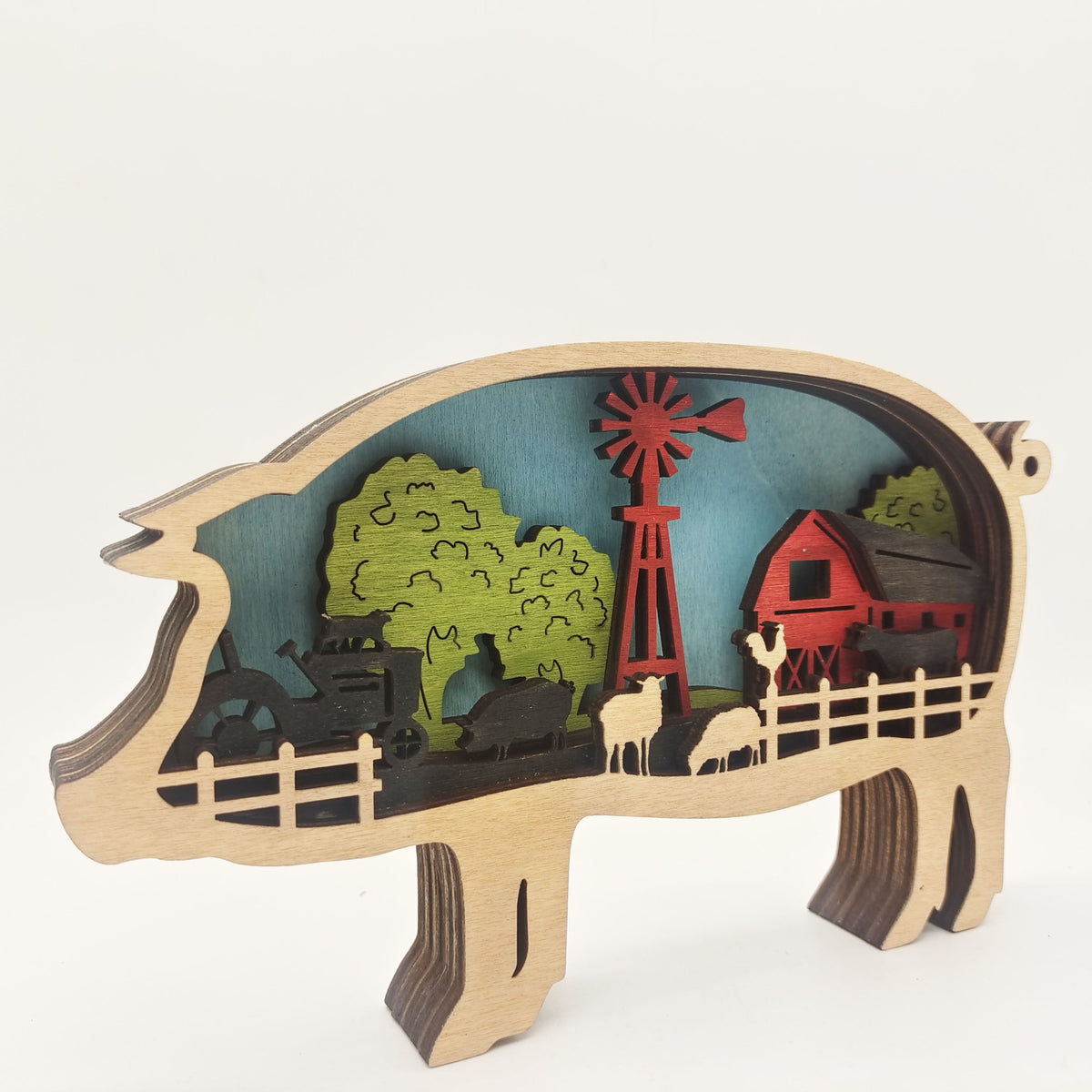 A wooden pig-shaped decorative sculpture featuring a layered farm scene with a red windmill, barn, trees, fence, and silhouettes of animals, photographed on a clean white background.