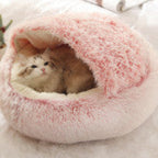A fluffy pink and white round pet bed with a cat curled up inside, set on a soft white surface with a small plant and magazine in the background.