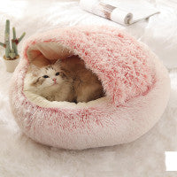 A fluffy pink and white round pet bed with a cat curled up inside, set on a soft white surface with a small plant and magazine in the background.