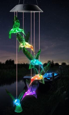 A hanging hummingbird wind spinner with multiple colorful bird figures illuminated in green, blue, orange, purple, and yellow, suspended under a black cup-shaped top against a nighttime outdoor scene near water.