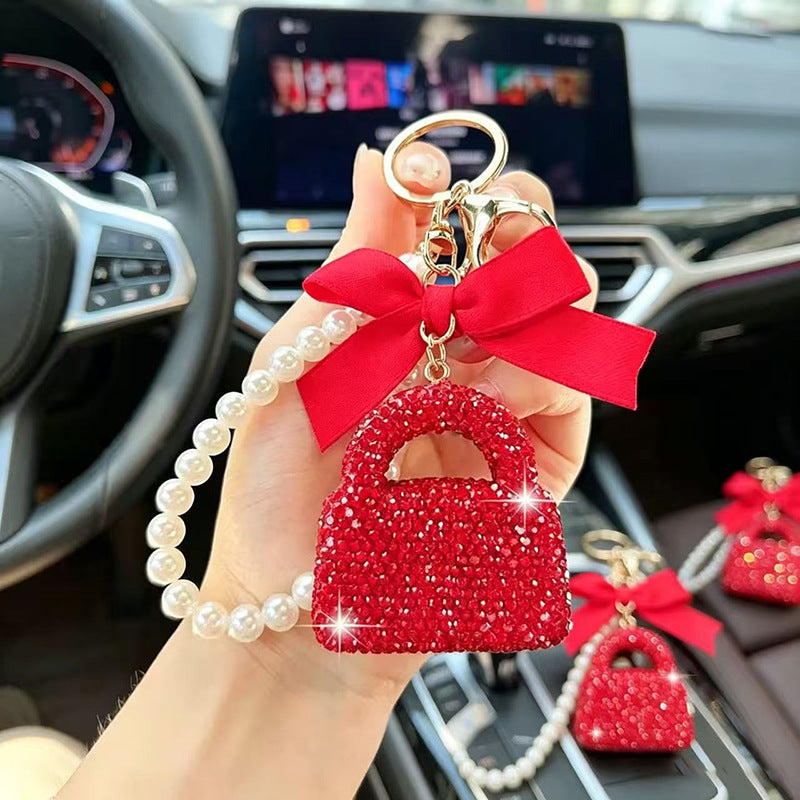 A hand holds a red rhinestone handbag-shaped keychain charm with a red ribbon bow and a pearl bead strap inside a car. Two matching charms are visible on the dashboard in the background.
