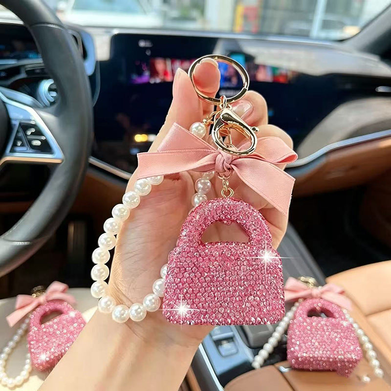 A hand holds a pink rhinestone mini handbag keychain with a pearl strap and pink ribbon bow inside a car. Two matching keychains are resting on the car seat in the background.