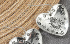 Silver heart-shaped dangle earrings with engraved sunburst and star details, displayed on a textured gray surface next to a woven straw mat.