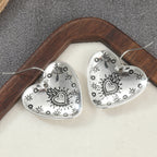 A pair of silver heart-shaped dangle earrings with engraved floral and heart motifs, displayed on a cream-colored surface with a dark wooden frame and gray textured background.