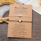 Two heart-themed cord bracelets displayed on a brown kraft card with printed sentimental text, photographed on a wooden surface with part of a gold mirror frame and a card visible in the background.
