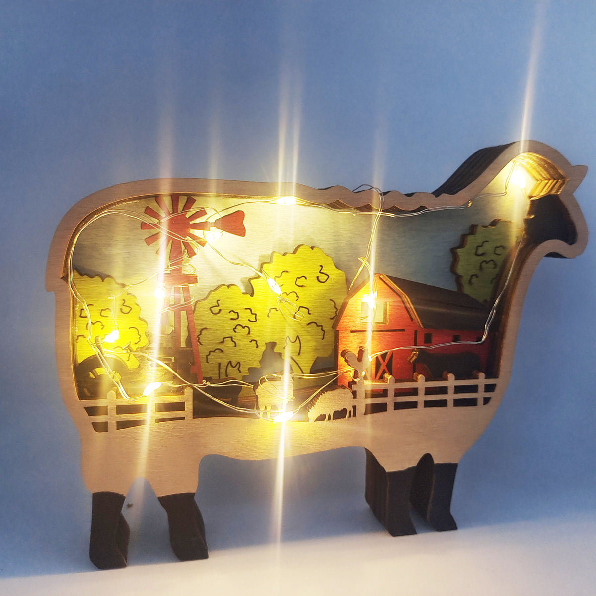 A wooden sheep-shaped decorative light featuring a farm scene with a red barn, yellow trees, a red windmill, and warm white string lights, shown against a blue gradient background.