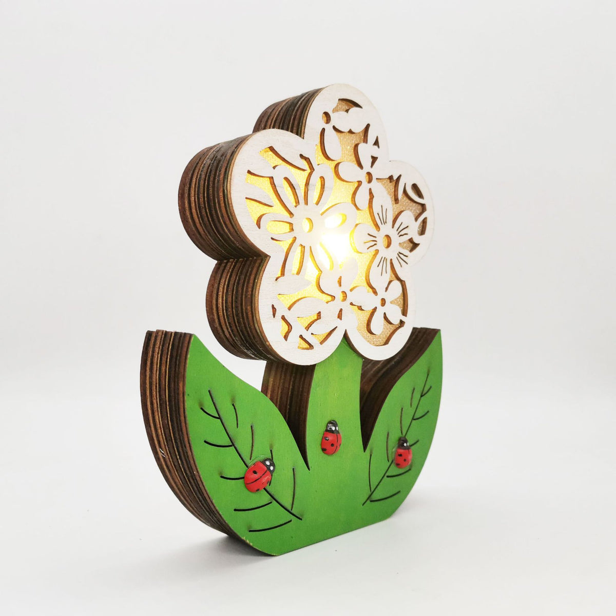 A decorative wooden flower-shaped light with a glowing warm interior, featuring layered wood petals, green leaf-like base pieces, and small painted ladybugs, photographed on a clean white background.