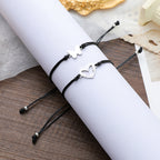 Two black cord bracelets with silver butterfly and heart charms, displayed on a white rolled-up sleeve over a wooden surface.