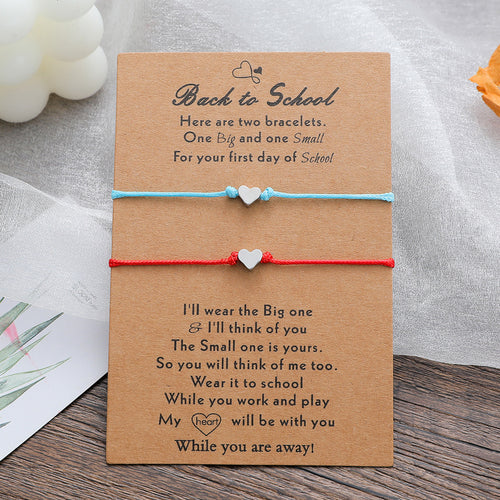 A brown kraft card with printed sentimental text, holding two heart charm string bracelets: one light blue and one red. The card is photographed on a wooden surface with soft fabric and decorative props in the background.