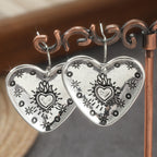 A pair of silver heart-shaped dangle earrings with black engraved floral and heart motifs, hanging from a copper-colored jewelry display stand.