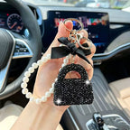 A hand holding a car keychain charm inside a vehicle. The charm features a black rhinestone-embellished purse-shaped pendant, a black satin bow, gold-tone key rings, and a white pearl bead loop.
