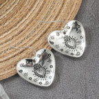 A pair of silver heart-shaped dangle earrings with engraved sunburst and star motifs, displayed on a textured gray surface next to a woven straw mat.