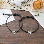 Two black cord bracelets with silver butterfly and heart charms, displayed on a dark wood surface with a lace doily, dried flower, and cards as props.