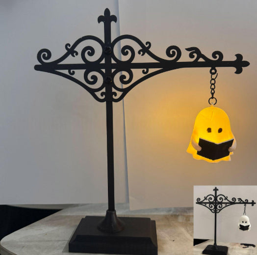 A black ornate metal display stand holding a glowing yellow ghost-shaped lamp that reads a black book. The main view shows the lamp lit against a light wall, with a smaller inset in the lower right showing the stand empty and the lamp unlit.