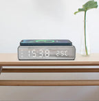 Digital clock with temperature display on a wooden surface, featuring a plant in the background.