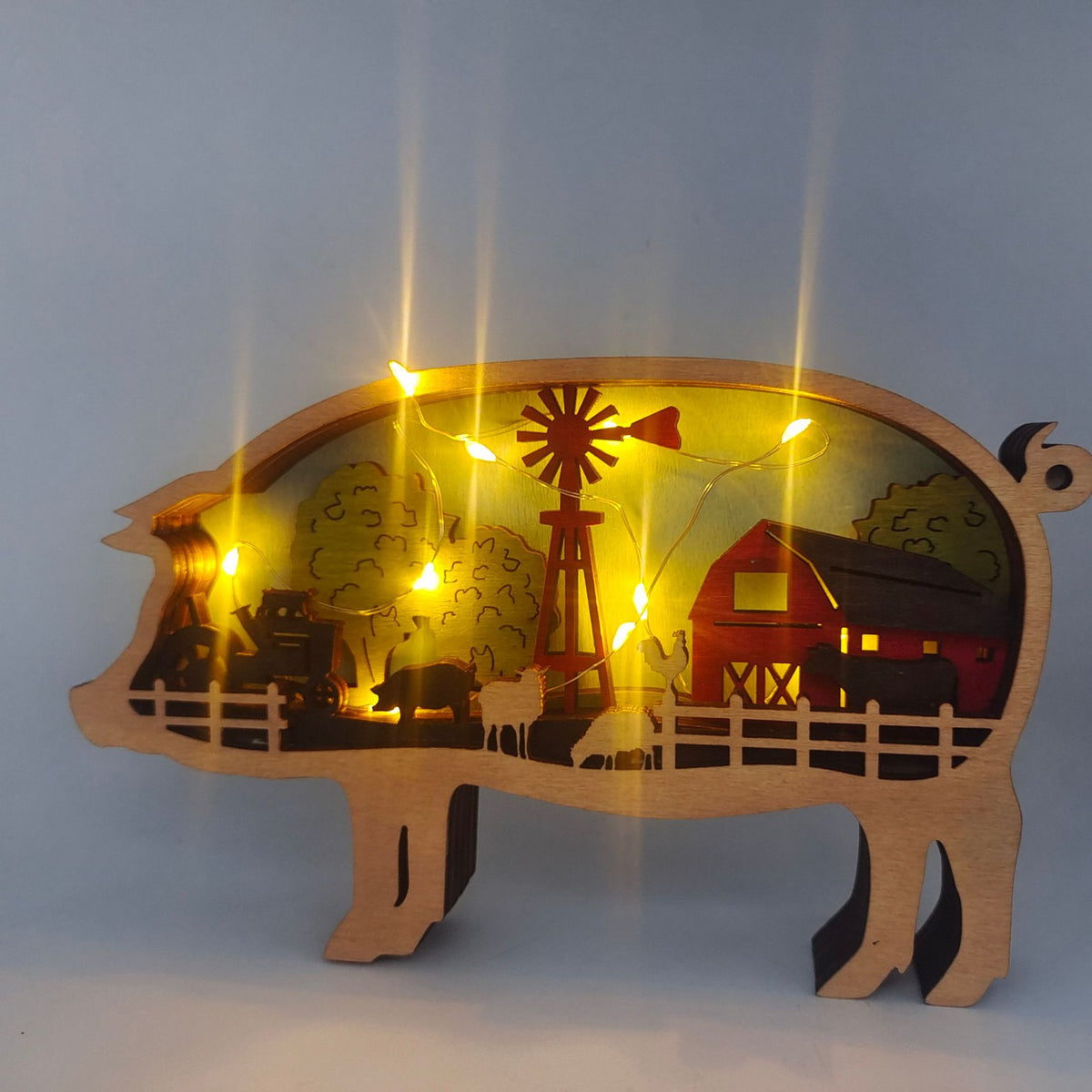 A wooden pig-shaped decorative light with a farm scene cutout inside, illuminated by warm yellow string lights. The silhouette shows a windmill, barn, fence, trees, and silhouettes of animals against a pale blue-gray background.