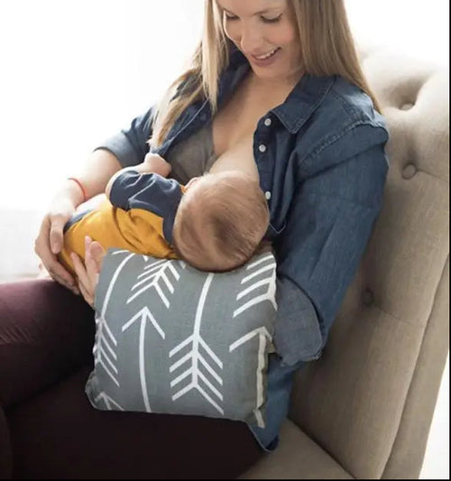 A woman in a denim shirt breastfeeds from a gray nursing pillow with white arrow patterns, seated on a beige armchair. Six additional pillow color and pattern options are shown in a vertical column on the left against a white background.