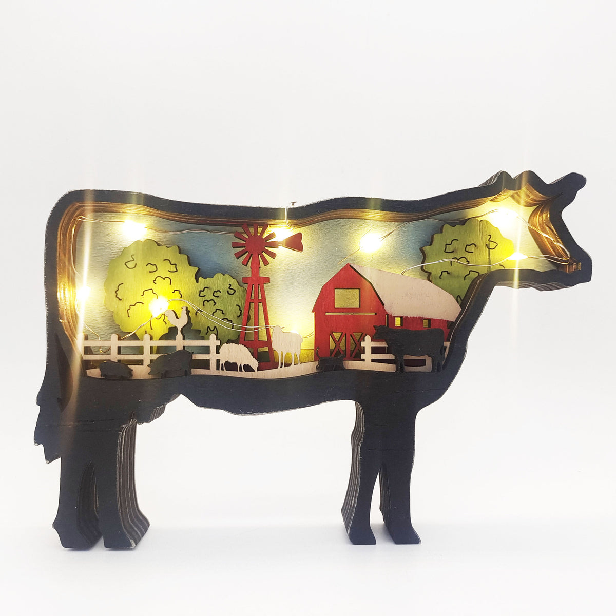 A cow-shaped decorative light fixture with a warm white LED interior showing a colorful farm scene with a red barn, trees, and windmill.