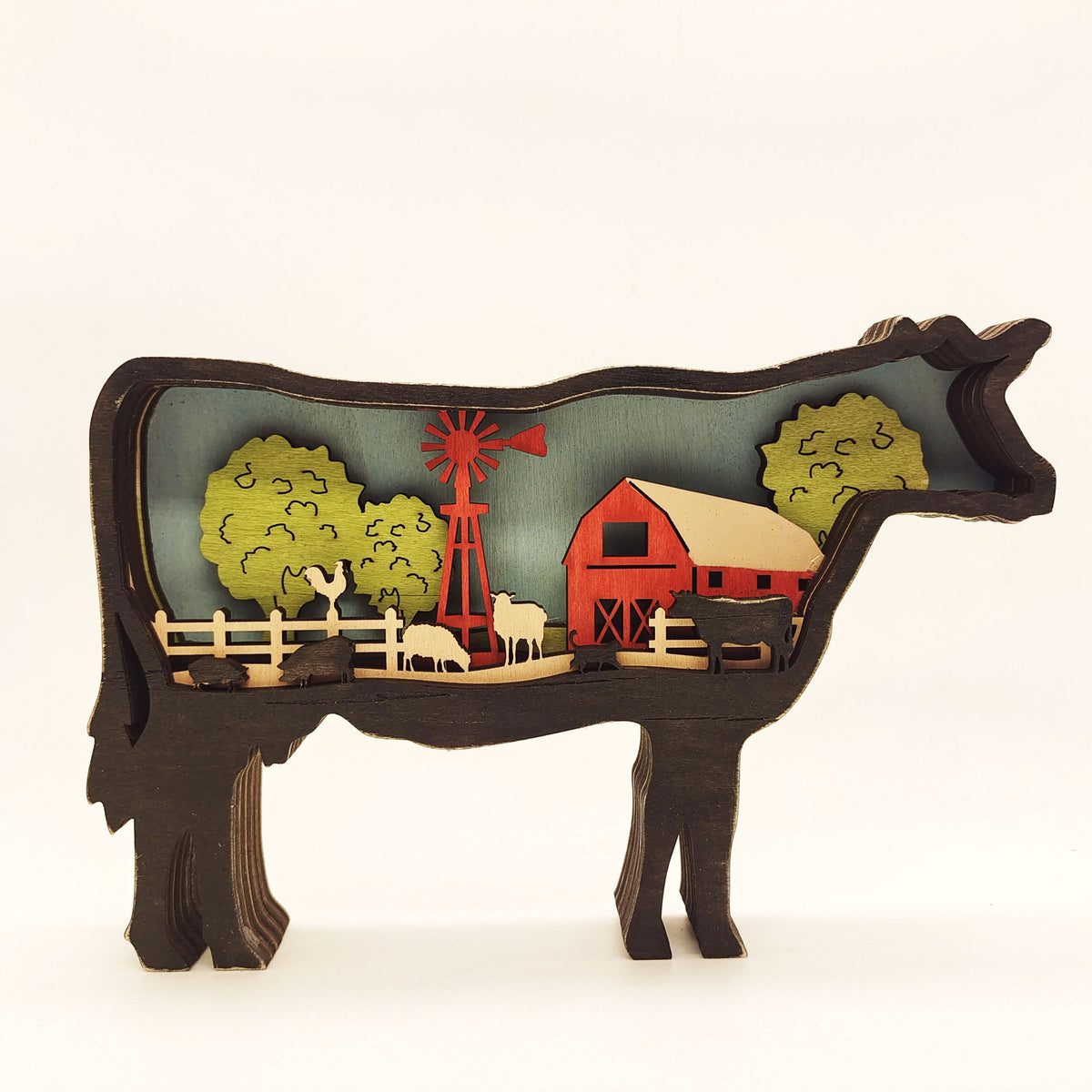 A decorative wooden cutout sculpture shaped like a cow, filled with a layered farm scene including a red barn, red windmill, green trees, white and black cows, and a light blue background, photographed on a clean white studio background.