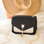 Black handbag with gold chain and cat-shaped charm on a soft surface with a straw hat in the background.