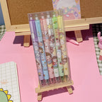 A set of six pastel-colored gel pens with cute cartoon characters printed on the barrels, packaged in a clear plastic sleeve and displayed on a small wooden stand against a pink desk background.
