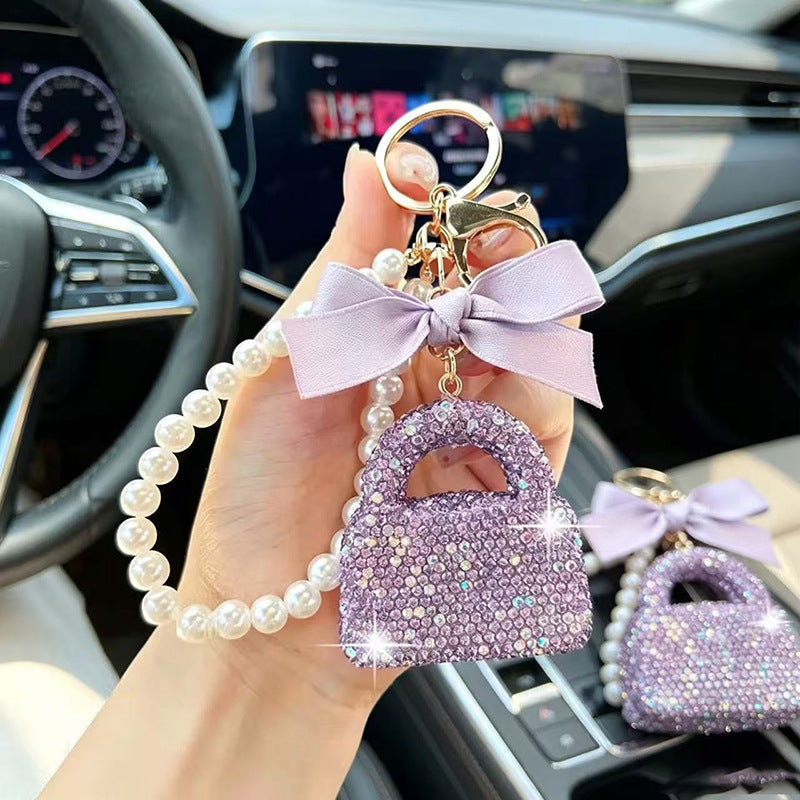 A hand holds a lavender rhinestone handbag-shaped keychain with a pearl strap and bow inside a car. Another matching keychain rests on the center console.