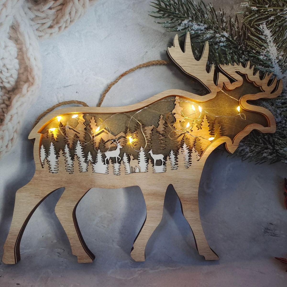 A wooden moose-shaped holiday decoration with a laser-cut forest and deer design, illuminated by warm string lights, displayed against a cozy winter background with pine branches and a knitted blanket.
