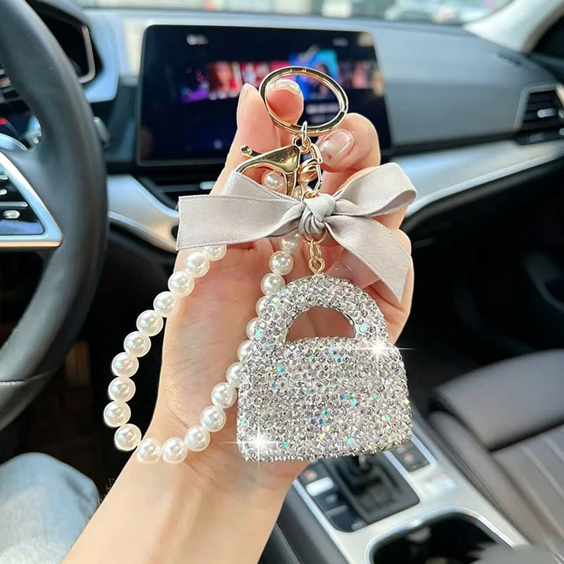 A hand holding a car keychain in a car interior. The keychain features a rhinestone-embellished padlock charm, a light beige satin bow, a pearl bead strap, and a gold-tone split ring.