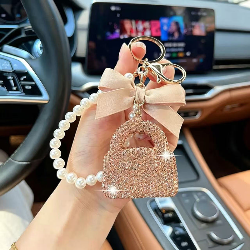 A hand holds a car keychain charm inside a car. The charm features a sparkling rose gold padlock, a pale pink satin bow, a pearl bead chain, and gold-tone metal rings.