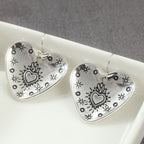 A pair of silver heart-shaped dangle earrings with black engraved floral and heart motifs, displayed on a white ceramic dish against a gray background.