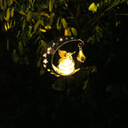 A glowing fairy-shaped solar garden light hanging from a tree branch at night, featuring a bright warm yellow light inside a crescent moon and star cutout frame.
