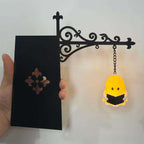 A hand holds a black decorative wall sign with an ornate scrollwork bracket and hanging chain. The sign has a rectangular black panel with a floral cutout and hangs next to a small yellow ghost-shaped light or ornament reading 'I AM A GHOST'.